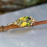 Close-up of a ring with an oval yellow-green gemstone and small diamonds on the sides, resting on a twig.