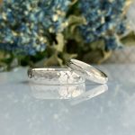Two hammered silver rings resting on a reflective surface with blue flowers blurred in the background