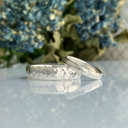 Two hammered silver rings resting on a reflective surface with blue flowers blurred in the background