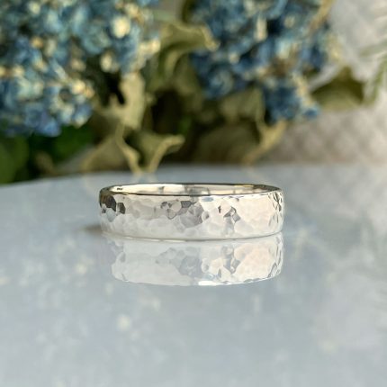 Hammered silver ring resting on a reflective white surface with blue flowers blurred in the background; a simple wedding/band style.