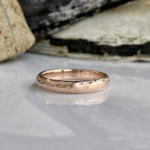 Rose gold textured wedding band resting on a reflective surface with rocks in the background