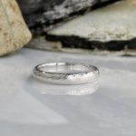 Silver textured wedding band resting on a reflective surface with rocks in the background, close-up view.