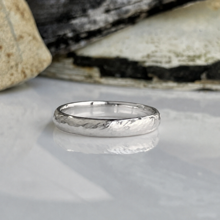 Silver textured wedding band resting on a reflective surface with rocks in the background, close-up view.
