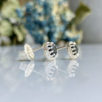 Silver stud earrings with an intricate scalloped lattice setting on a reflective surface, blurred blue flowers in the background.