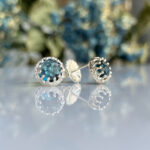 Round blue-green gemstone stud earrings in a silver setting on a reflective surface with a blurred floral background.