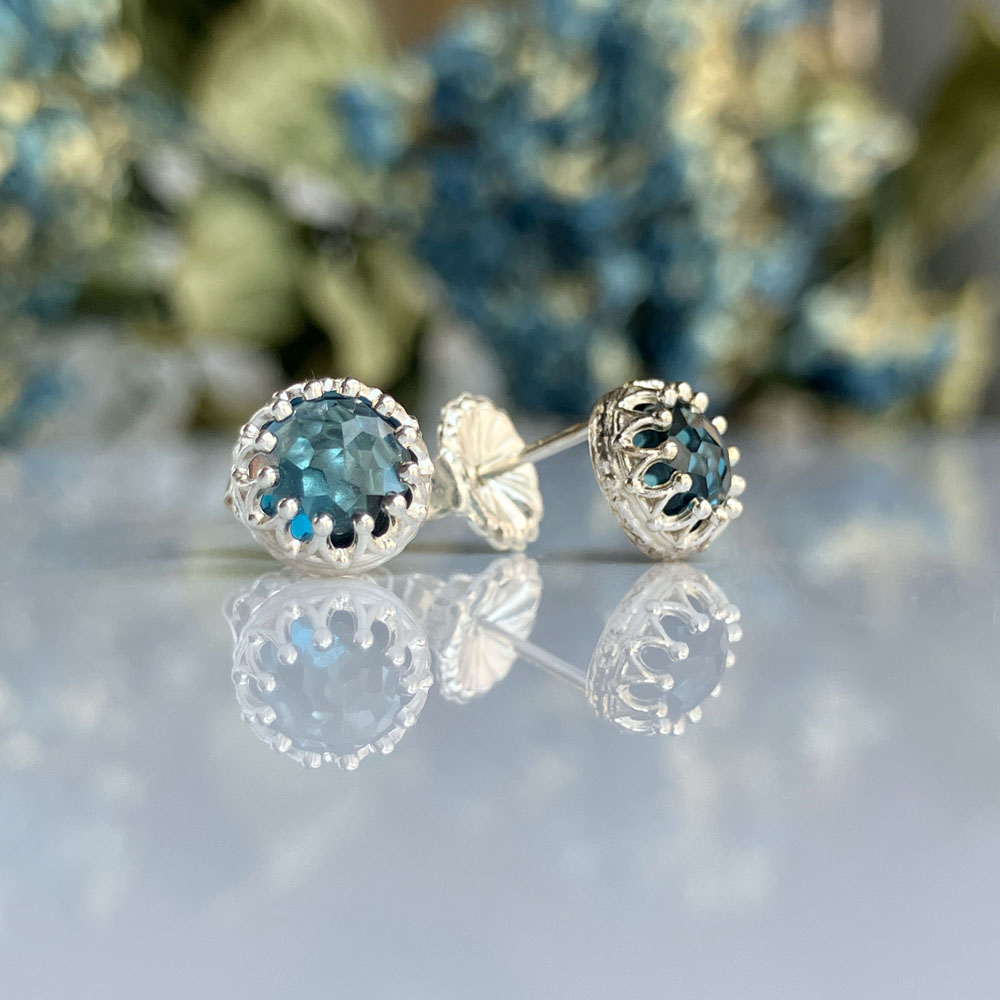 blue topaz silver crown setting earringsjpeg | Posy Fine Jewellery Toronto Round blue-green gemstone stud earrings in a silver setting on a reflective surface with a blurred floral background.