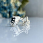 Silver stud earring with a black gemstone and intricate filigree, placed on a reflective surface.