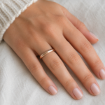 Close-up of a left hand with a rose-gold wedding band on the ring finger, resting on a light surface.