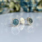 Stud earrings with blue-green gemstones in silver scalloped settings on a reflective surface.