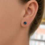 Close-up of a woman's ear wearing a round blue gemstone stud earring with a gold beaded halo.