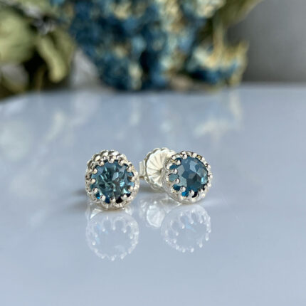 Silver stud earrings with round blue-green faceted stones in a scalloped bezel, placed on a reflective surface with blurred floral background
