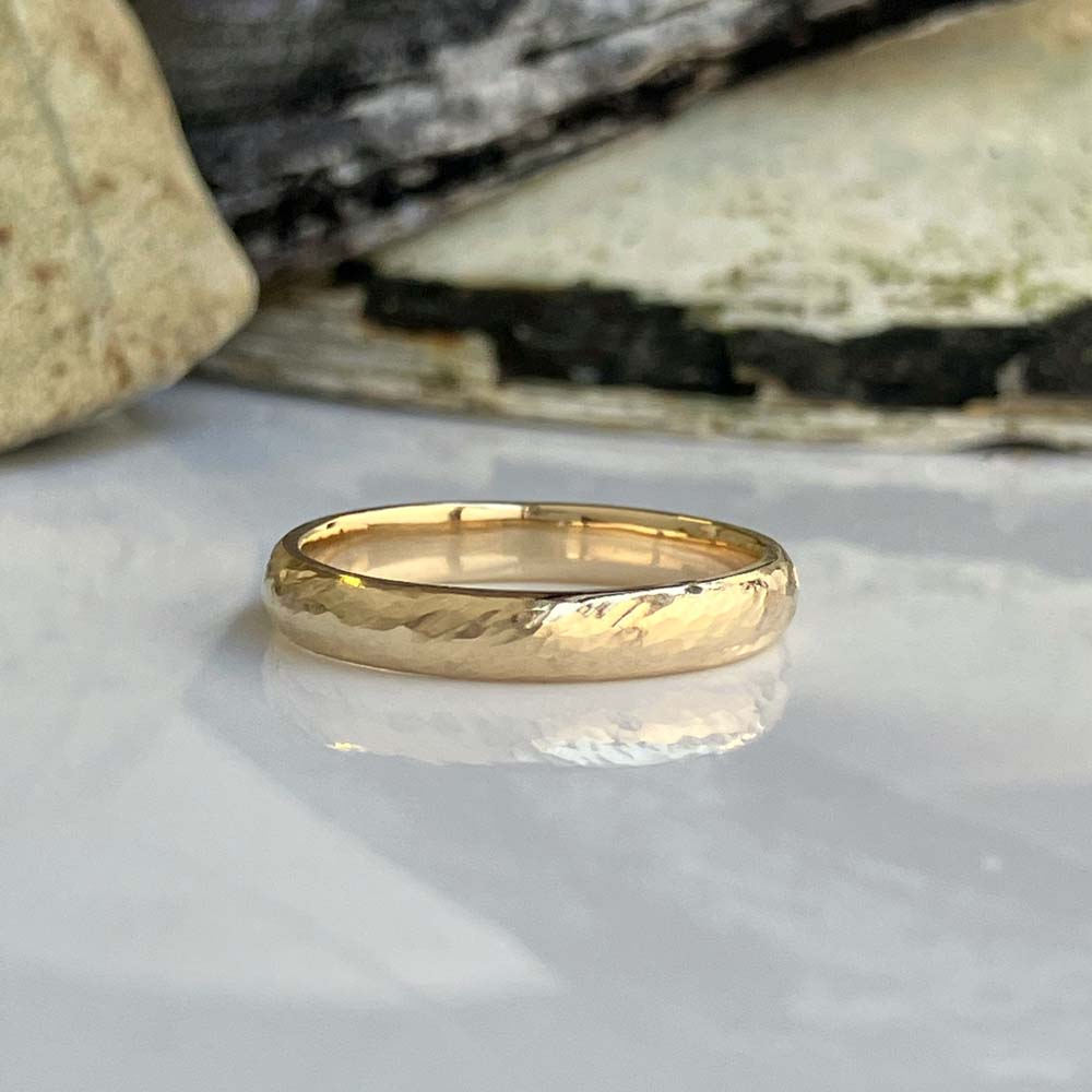 waves at the shore ladies wedding bandjpeg | Posy Fine Jewellery Toronto Gold wedding band resting on a glossy white surface with a stone backdrop behind it.