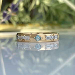 Gold ring with a square blue gemstone center and small side diamonds, photographed on a reflective surface with a blurred floral background.