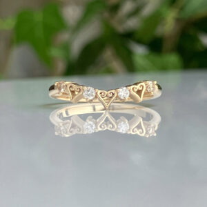 Gold ring with heart motifs and small diamonds, reflected on a glossy surface.