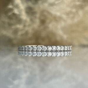 Thin diamond eternity ring with a continuous row of round stones in a white metal band, reflected on a mirrored surface.