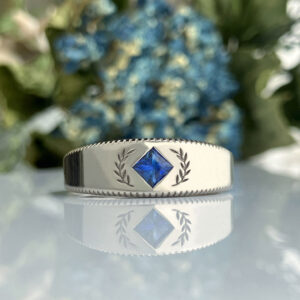 Silver ring with a blue square gemstone centered between laurel motifs, reflected on a glassy surface.