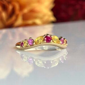 Gold ring with five gemstones—pink, yellow, pink, yellow, pink—centered pink gem, on a reflective surface.