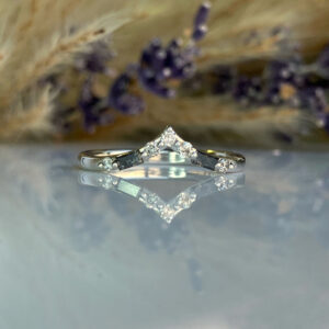 Diamond V-shaped ring with a central peak, reflected on a glossy surface with blurred dried flowers in the background.