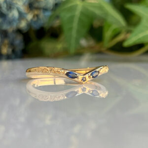 Gold ring with blue gemstones and floral engravings, resting on a reflective surface with greenery in the background