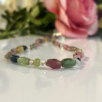 Multicolored gemstone bracelet on a white reflective surface, with a pink rose blurred in the background.