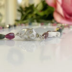 Close-up of a silver bead bracelet with a lattice-cut bead and purple and green faceted stones on a reflective white surface; a small engraved tag and blurred pink flowers in the background.