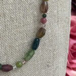 Beaded necklace with multicolored stones (green, teal, pink, purple) draped on beige burlap fabric.