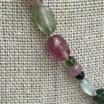 Close-up of a beaded necklace with faceted green, purple, and pink stones on a burlap background.