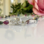 Close-up of a beaded silver bracelet withpurple and green stones and an ornate clasp, reflecting on the surface.