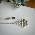 Silver necklace featuring a hexagonal honeycomb pendant on a delicate chain, set against a white surface with dried blue flowers nearby.