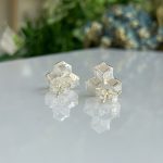 Pair of silver cube-stud earrings with a small bow, placed on a reflective surface with a blurred green background.