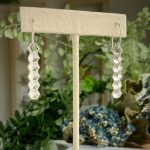 Dangling silver earrings with stacked hexagonal beads displayed on a beige jewelry stand.