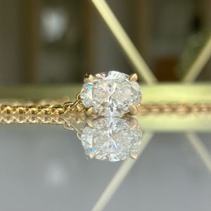 Gold chain with a round brilliant-cut diamond pendant in a four-prong setting, resting on a reflective surface with its reflection visible.