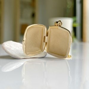 Open gold square locket with a chain resting on a white seashell, reflected on a glossy surface.