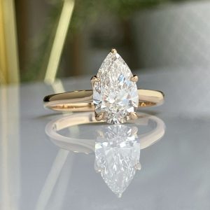 Pear-shaped diamond engagement ring on a gold band, shown with a reflective surface beneath it.