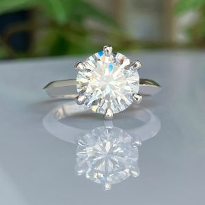 Diamond solitaire ring with a round-cut gemstone on a silver band, reflected on a glossy surface.