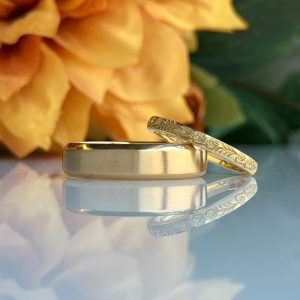 Two gold wedding bands, one plain and one engraved, rest on a reflective surface with a yellow flower backdrop.
