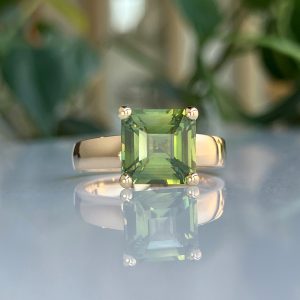Emerald-cut green gemstone on a reflective surface with small metal corner fittings and a blurred leafy background.