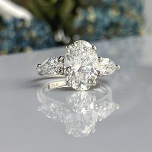Diamond ring with a large oval center stone and two pear-cut side stones on a reflective surface; platinum setting with 14K stamp visible on the band.
