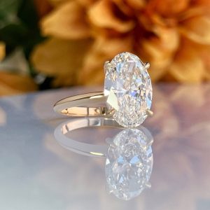 Diamond engagement ring with a large oval diamond on a gold band, reflected on a glossy surface with blurred orange flowers in the background.