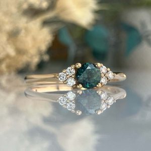 Gold engagement ring with a dark teal center gemstone surrounded by small diamonds, set on a slim band, reflecting on a glassy surface.