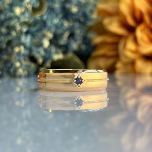 Gold ring with a blue gemstone set in the center, reflected on a glossy surface with a soft blurred background