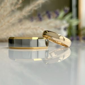 Two gold rings, one plain band and one with a faceted gray stone, resting on a reflective surface with a soft background.
