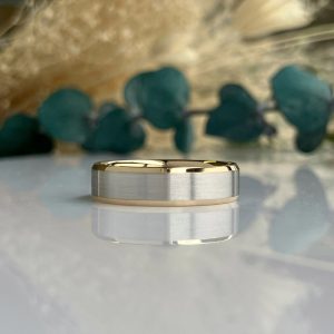 Two-tone wedding band with brushed silver center and gold edges on a glossy surface, blurred green background.