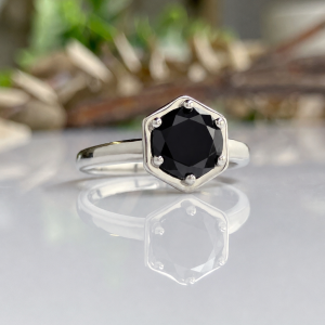 Silver ring with a dark, faceted hexagonal gemstone held by six prongs, reflected on a glossy surface.