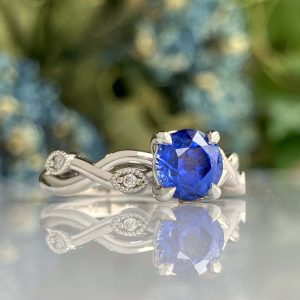 Silver ring with a cushion-cut blue sapphire centerpiece and a twisted band adorned with small diamonds, reflected on a glossy surface.