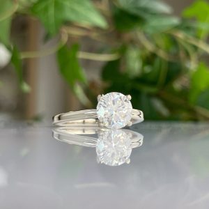 Diamond engagement ring with a round-cut diamond on a silver band, reflected on a glossy surface with green foliage in the background.