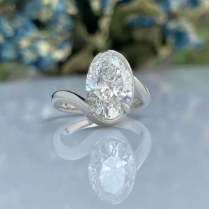 Close-up of an oval-cut diamond engagement ring with a sleek silver band, resting on a reflective surface with a soft blue floral background.