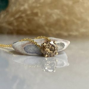 Gold necklace with a round faceted brown gemstone resting on a smooth stone, reflected on a glossy surface