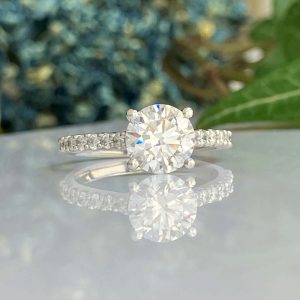 Engagement ring with a round-cut center diamond and pavé band, resting on a reflective surface.