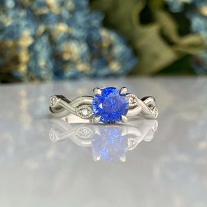 Silver ring featuring a vibrant blue faceted sapphire, set in a twisted band with small diamond accents, reflected on a glossy surface.